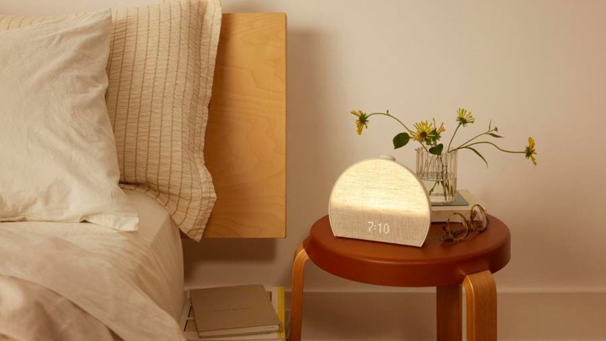 Hatch Restore 3 sunrise alarm clock on wooden bedside table alongside yellow flowers and a pair of glasses next to bed with beige bedding.