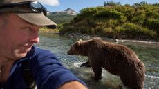 A photographer takes a selfie as a brown bear walks past.