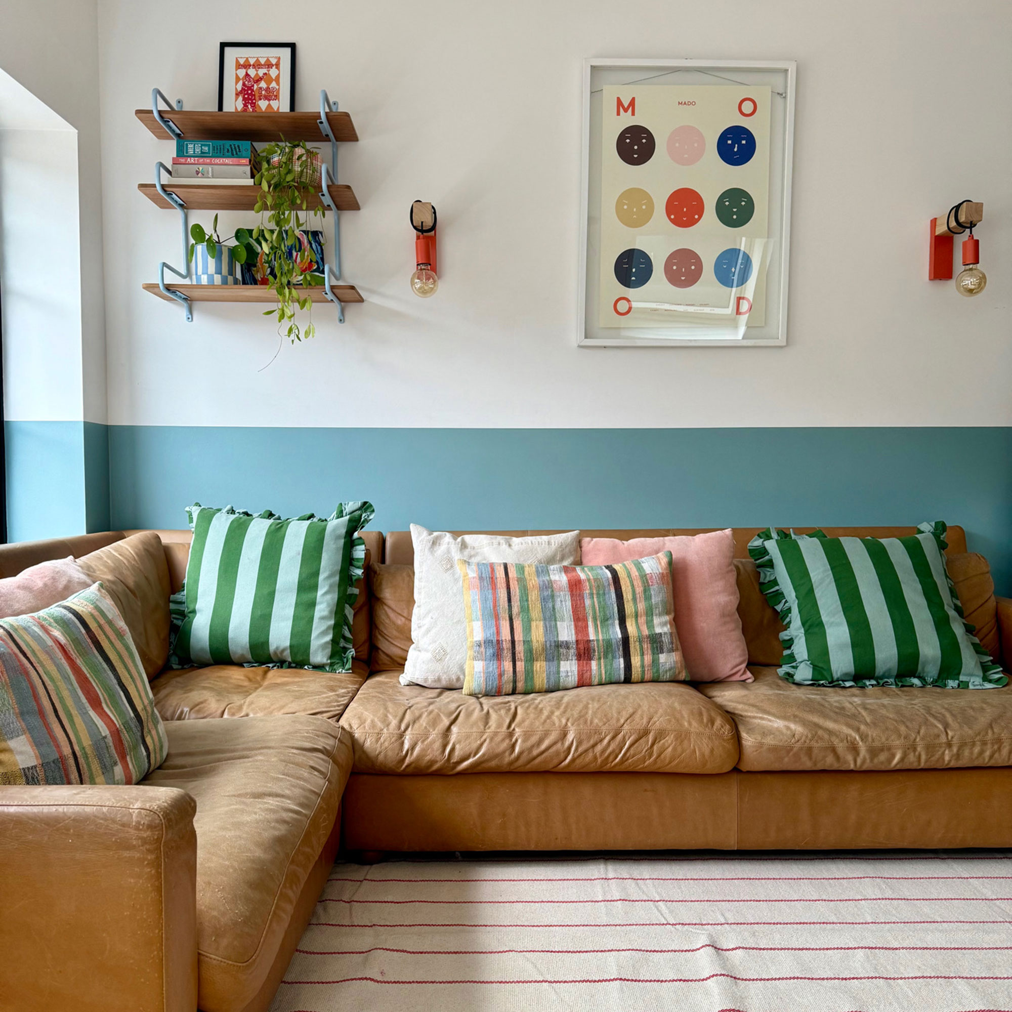 Living room with lower half of wall painted blue and upper white, low tan leather L-shaped sofa and striped colourful cushions