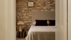 A stone bedroom wall with a hanging woven pendant lamp, a low gray bed with a pair of purple pillows, beside a low wood stool with a woven lamp and a vase