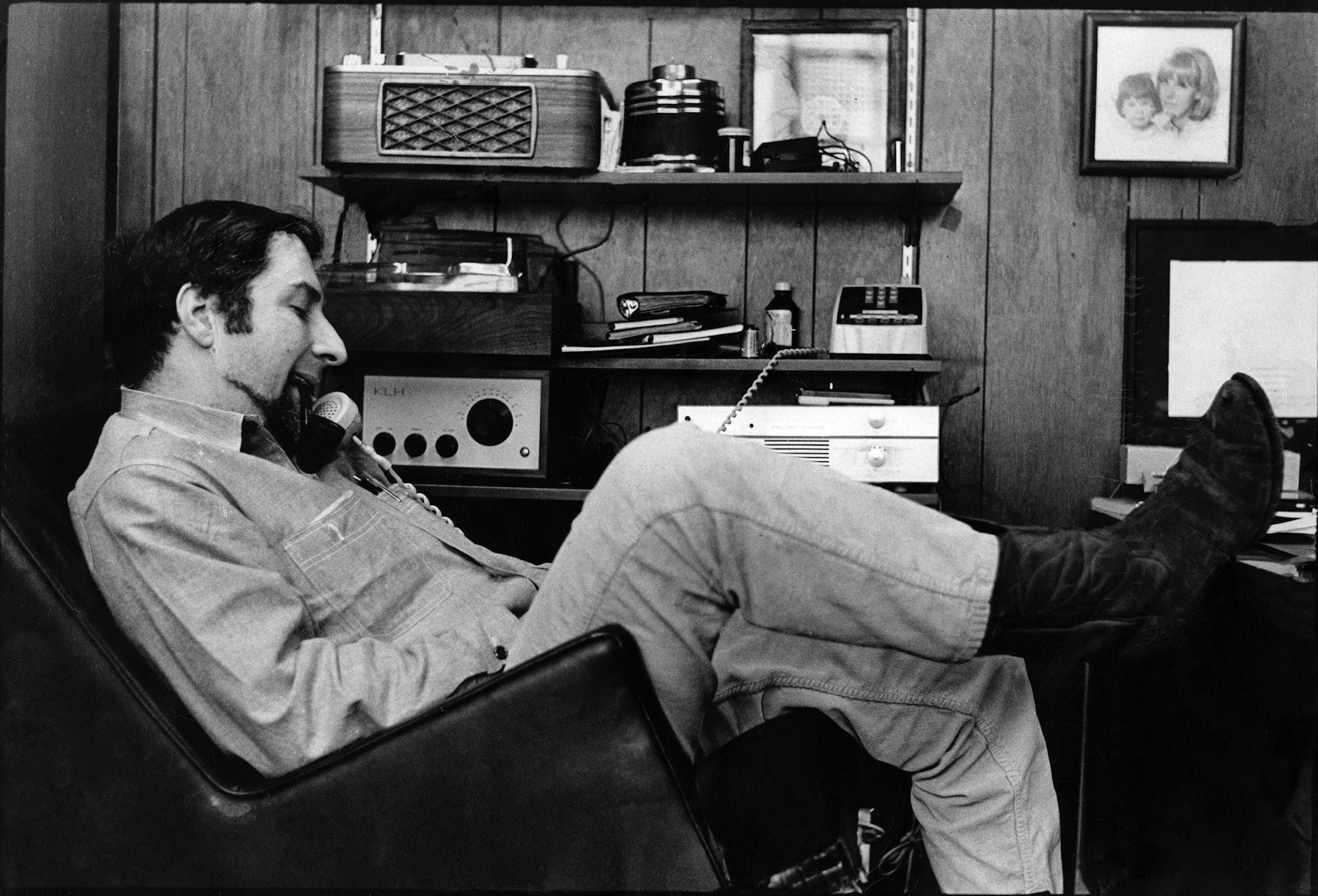 A black and white self-portrait of photographer Don Bronstein reclining in a chair with his feet up in his Chicago studio.