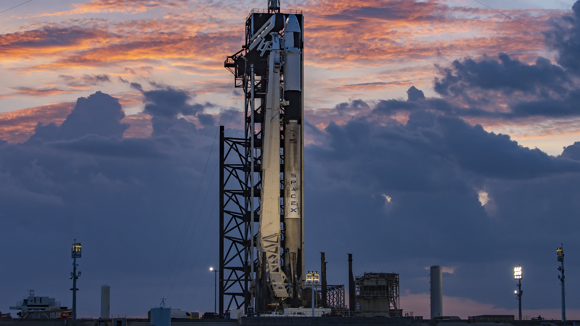 A black and white SpaceX Falcon 9 rocket carrying the Ax-4 crew capsule on the launch pad with colorful clouds lit by twilight.