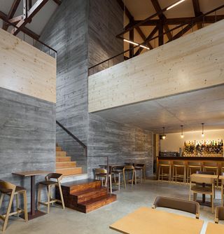 An interior view of the two-floor Entre Portas restaurant. Floors, walls, and ceilings are all in gray concrete style. The bar brown color is on the far wall, and we see bottles of drinks behind it. The tables and chairs are all in light wood. To the left, there are wooden stairs that lead to the second floor.