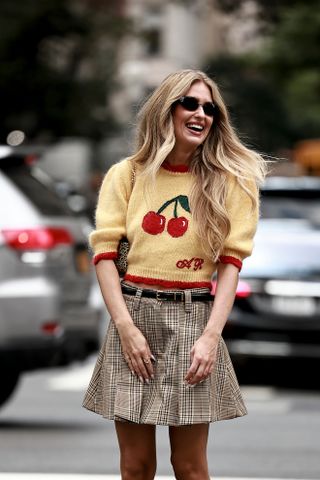 A woman wearing a yellow cherry short-sleeve sweater, black belt, plaid mini skirt, and black sungalsses.