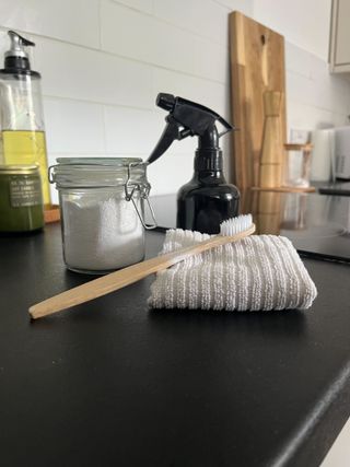 A brown spray bottle of vinegar next to a glass container of oxygen bleach. A microfiber cloth and toothbrush in front of them