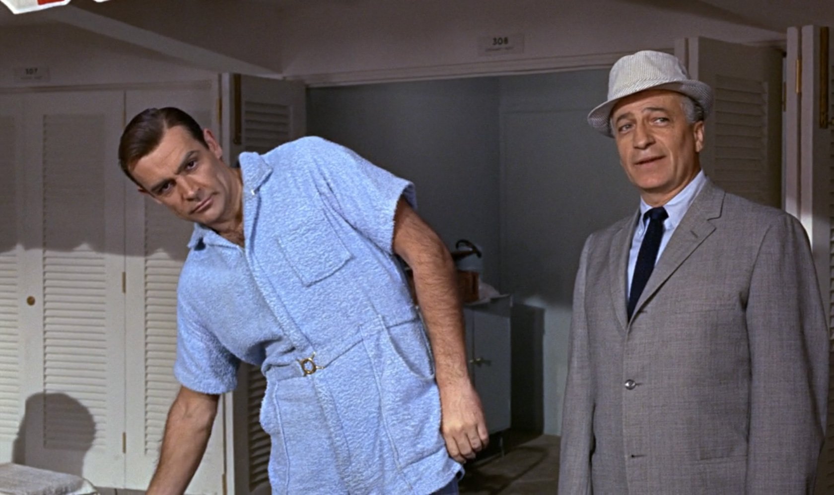Sean Connery in a baby blue terry towelling romper suit in a still from the Goldfinger film, standing next to another actor in a grey suit, blue shirt, navy blue tie and white Panama hat