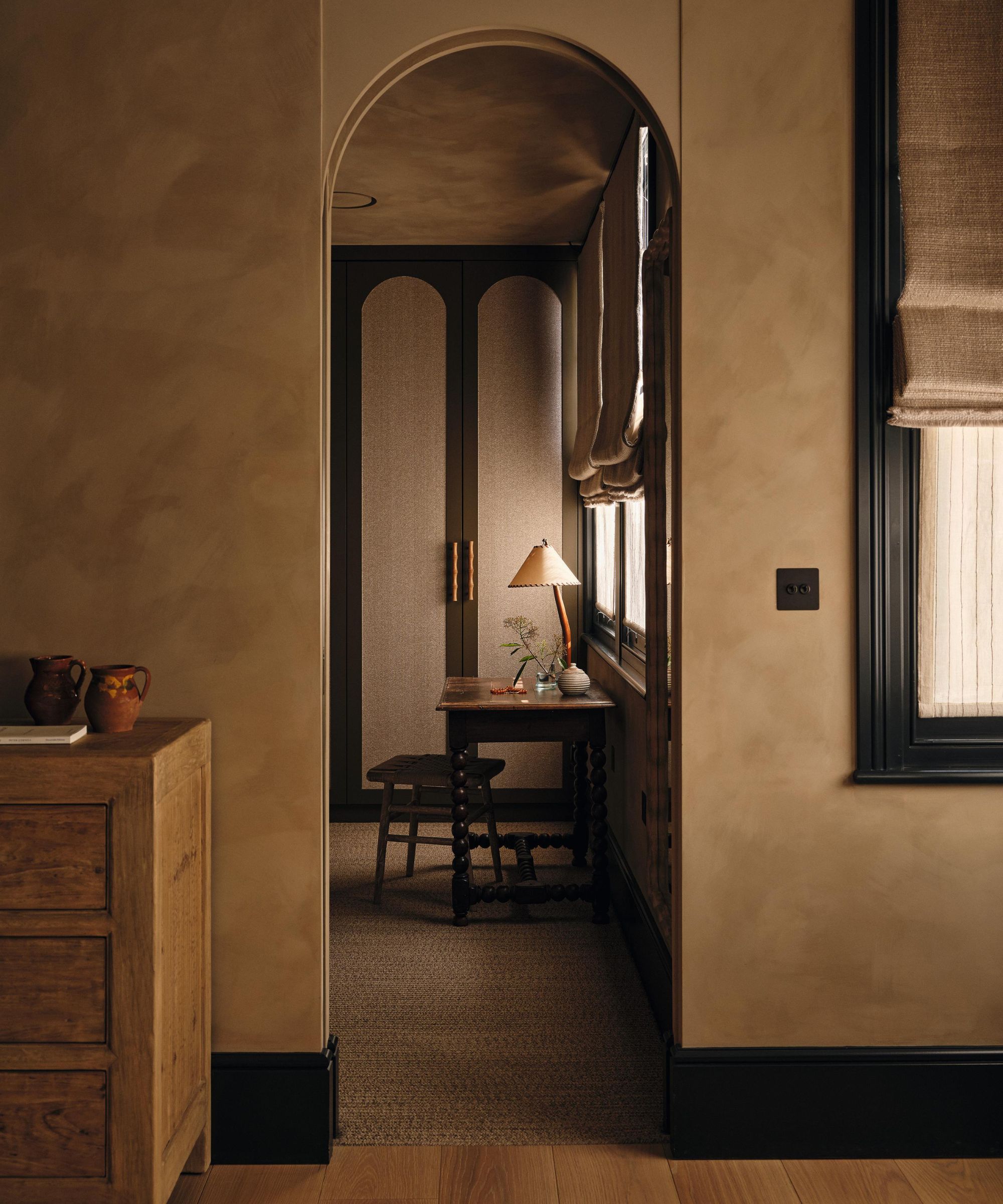 a beige limewashed bedroom with dark painted wood trims with an arched doorway through to a dressing room