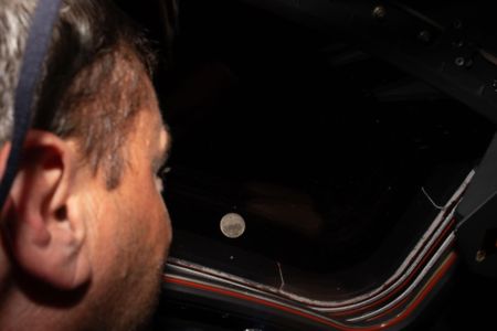 A man looks out a window. The moon is exceptionally clear in the background.