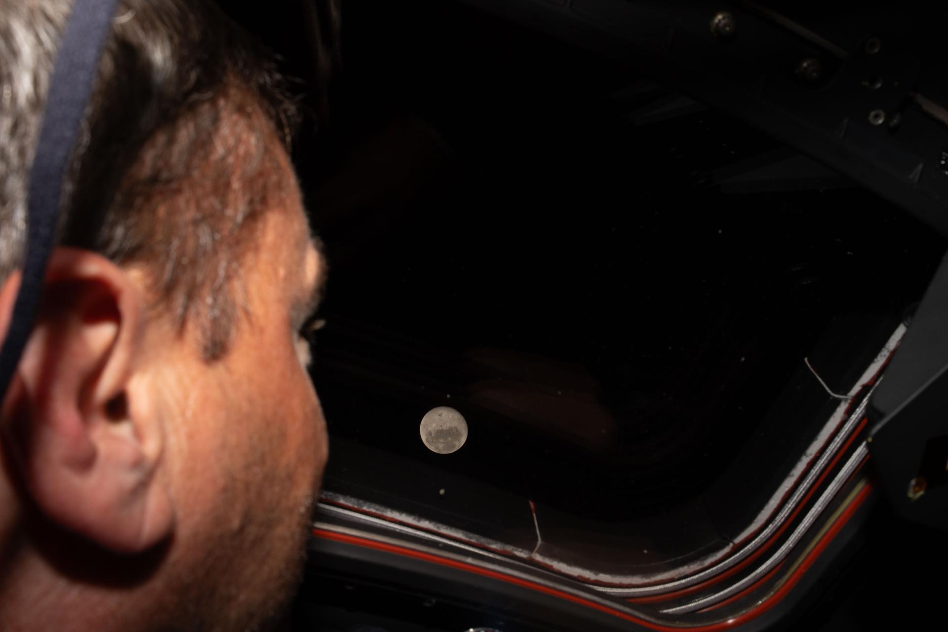 A man looks out a window. The moon is exceptionally clear in the background.