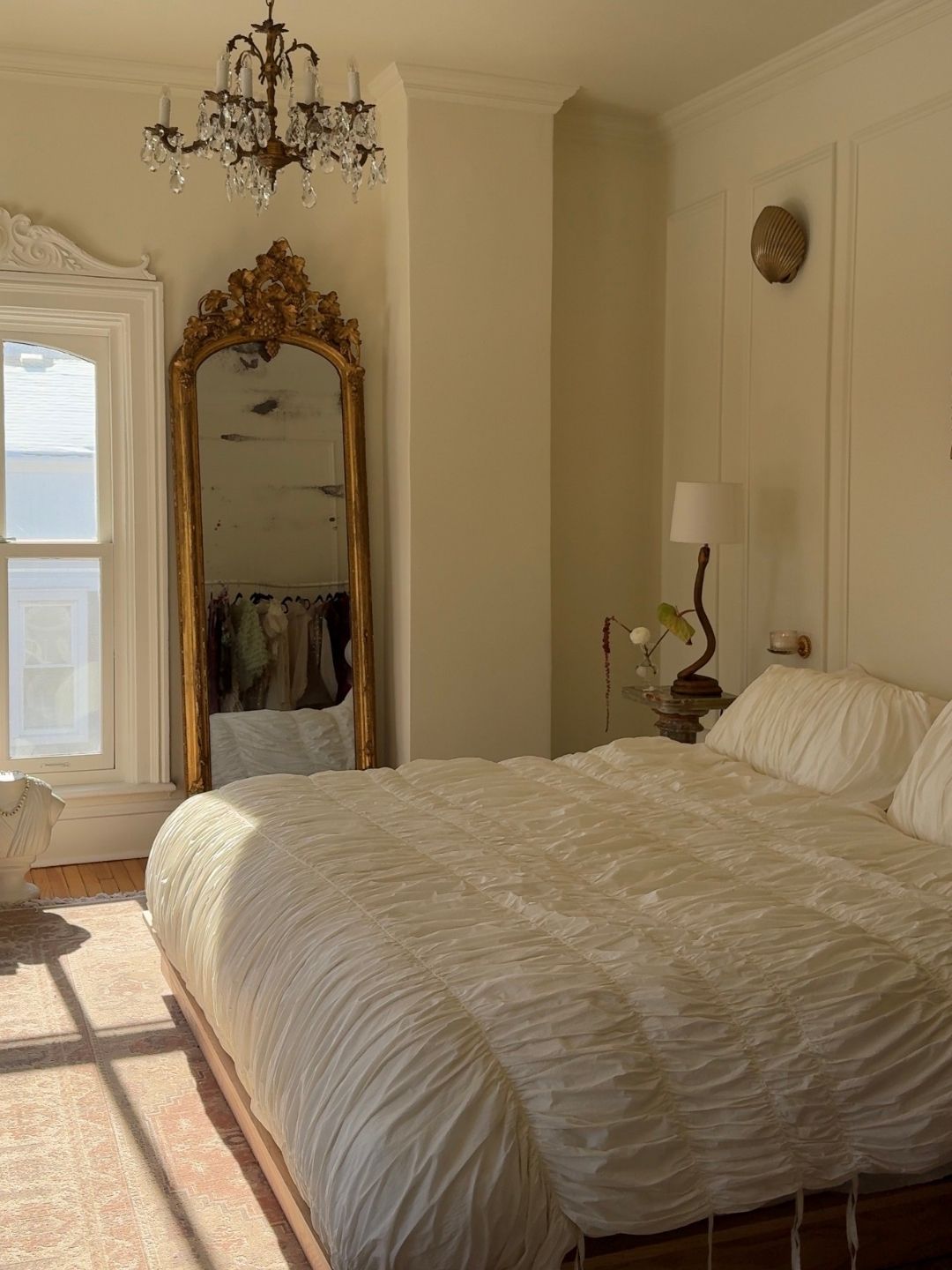 Image of a vintage-style white bedroom with ruched bedding, a floor-length gold vintage mirror, a chandelier, and a lamp by the bed.
