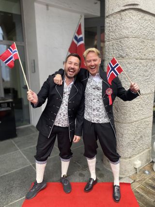 Martin Compston and Phil MacHugh outside a building wearing Norwegian national dress: black blazers and trousers, paisley waistcoats and knee socks. They are both waving Norwegian flags and Phil has his arm around Martin's shoulder.