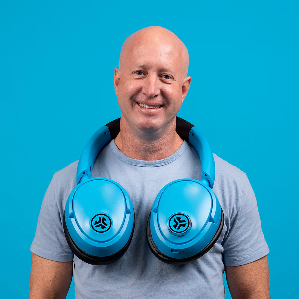 A man wearing the massively oversized JLab Blue XL Speaker Headphones around his neck in front of a blue background.