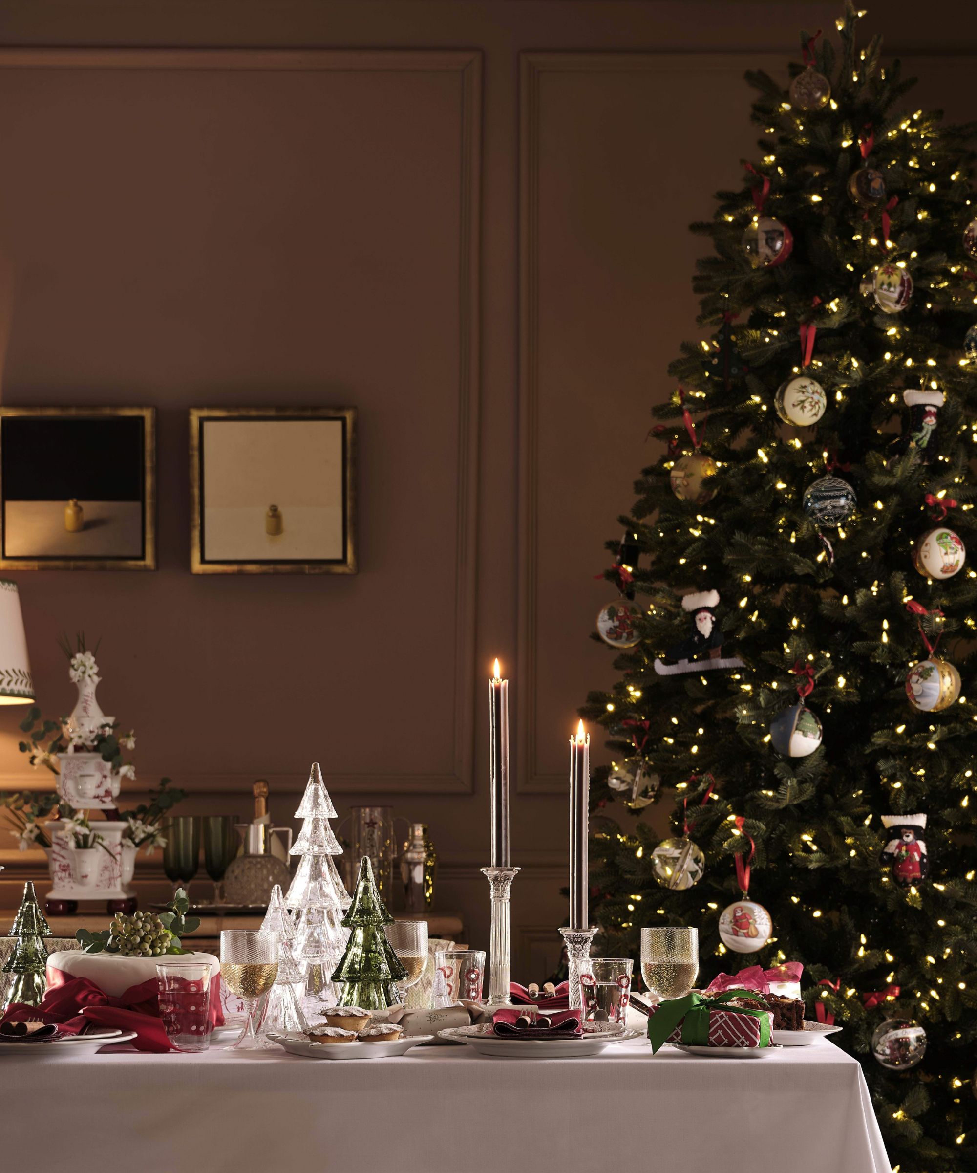 A dining table with a white tablecloth filled with festive decor, including candles, mini Christmas trees, and place settings. In the background, there is a large Christmas tree with lights and decorations.