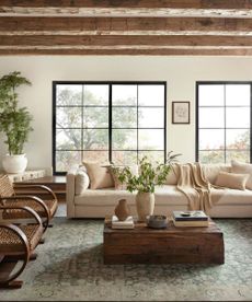 A cream sofa with matching throw pillows behind a wooden coffee table, covered with vases and books; to the left, wooden armchairs; in the background, windows looking onto a garden view.