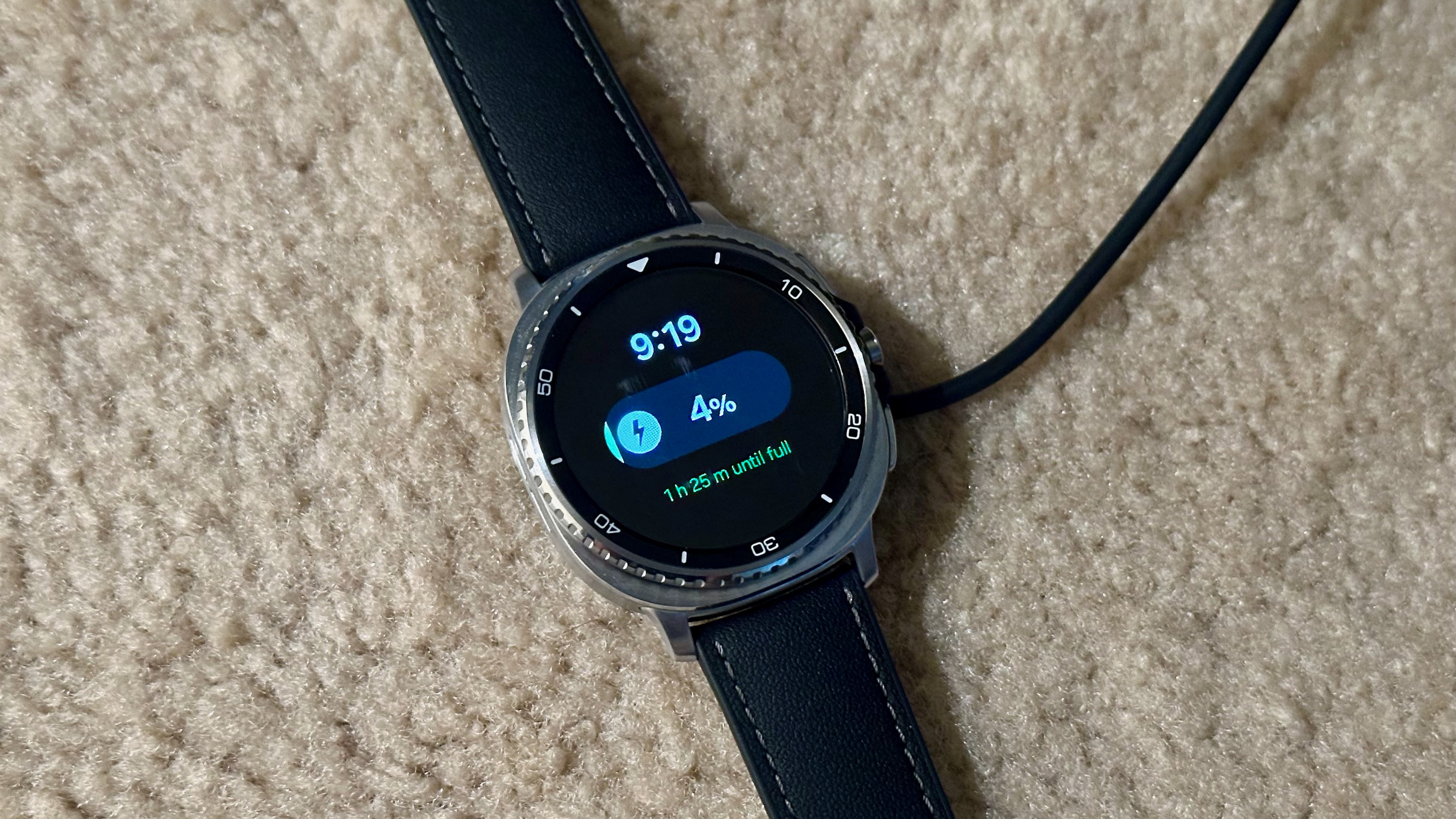 The Samsung Galaxy Watch 8 Classic atop its charger on carpet, the display showing 4% battery and 1 hr 26 minutes until full.