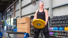 Woman in gym holds yellow weight plate in both hands. She is looking to the side and her mouth is open, smiling. She wears black pants and a black tank top. She has short grey hair with a tinge of purple dye. 