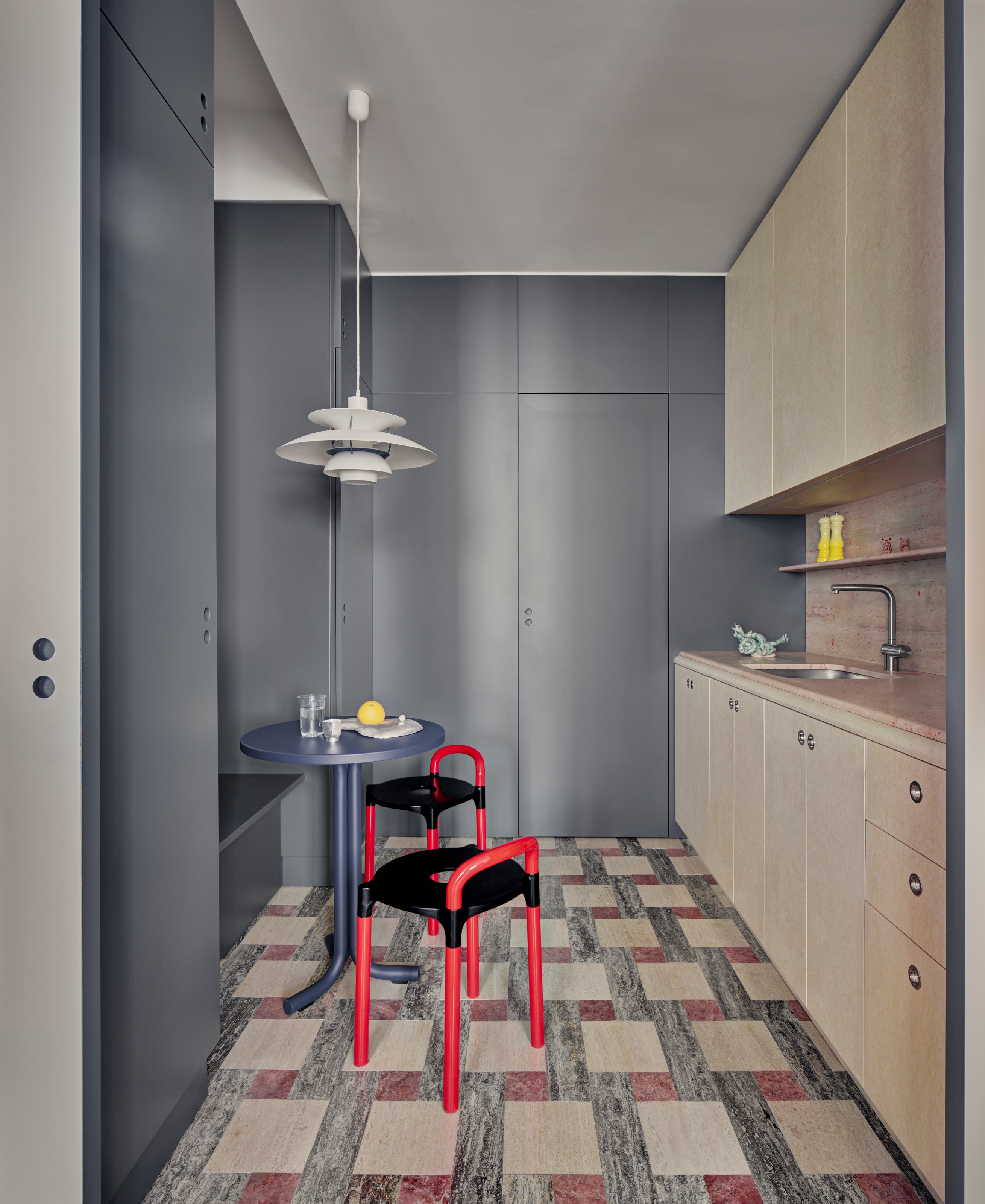 Kitchen with cream units, pink marble backsplash, powder blue storage on opposite side with built-in bench, small blue breakfast table and red and black chairs