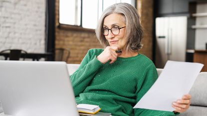 An older woman looks thoughtful as she looks at her laptop and holds paperwork.