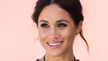 Meghan Markle wearing a black dress with her hair up in front of a plain backdrop