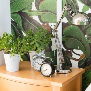 Black and silver alarm clocks next to silver lamp and plants on wooden bedside table