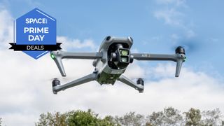 A Grey quadcopter with a black camera lens on the front is hovering in front of blue skies and the top of a tree line with the space.com logo in the top left corner.