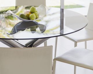 Round glass dining table with white chairs around and fruit bowl in the center