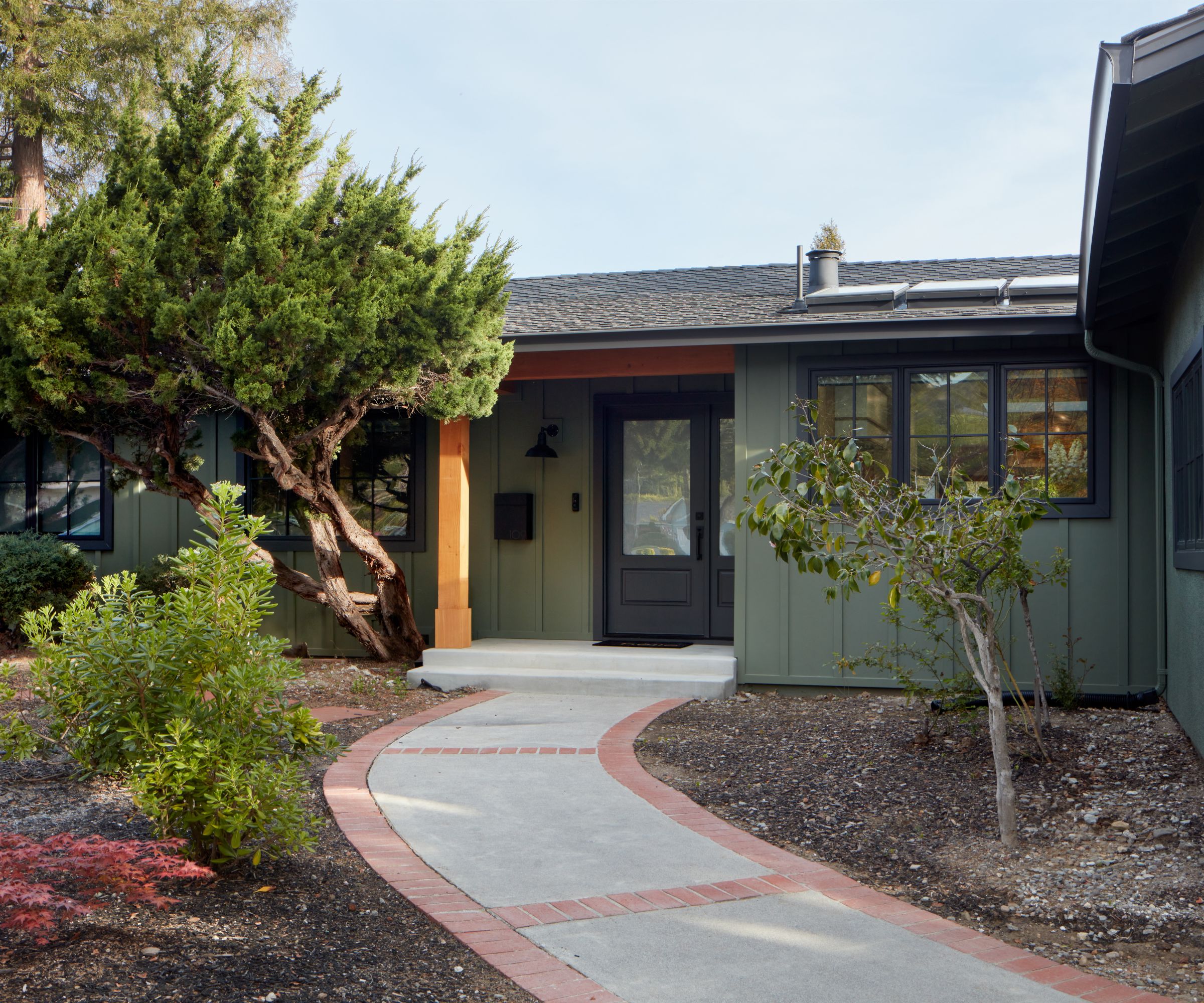 A bungalow home exterior with dark green siding, a charcoal gray front door and window trim, and an orange pillar. A paved pathway with surrounding plants.
