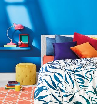 A bed furnished with bold assorted coloured cushions with a mustard yellow pouffe against a blue background