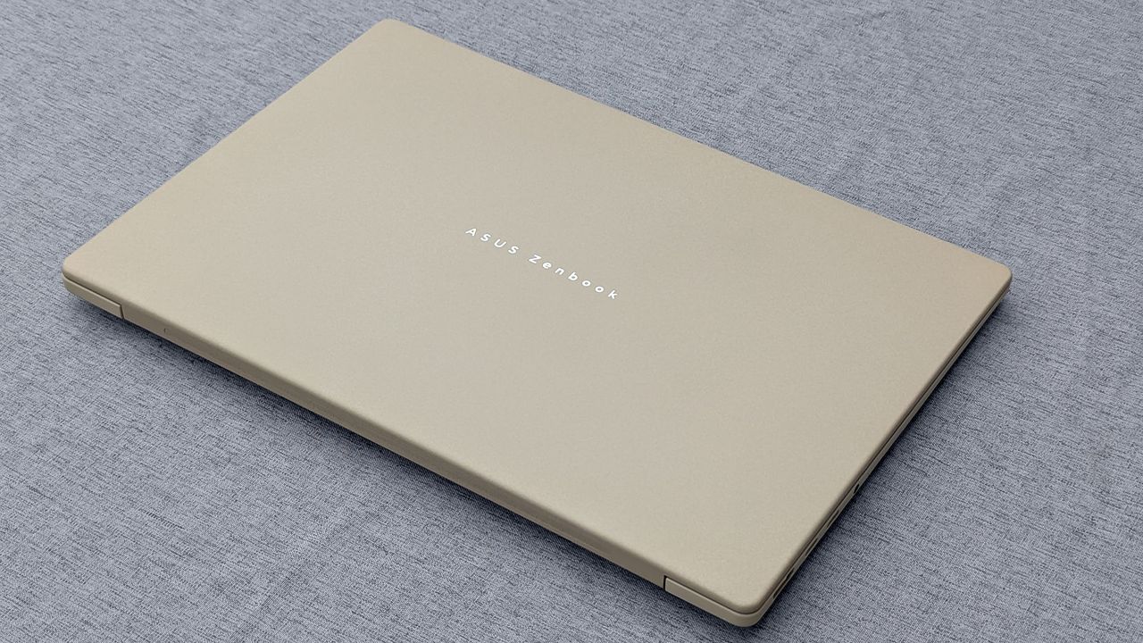 Closed beige ASUS Zenbook A14 resting on gray fabric, highlighting its slim metallic lid and simple branding.