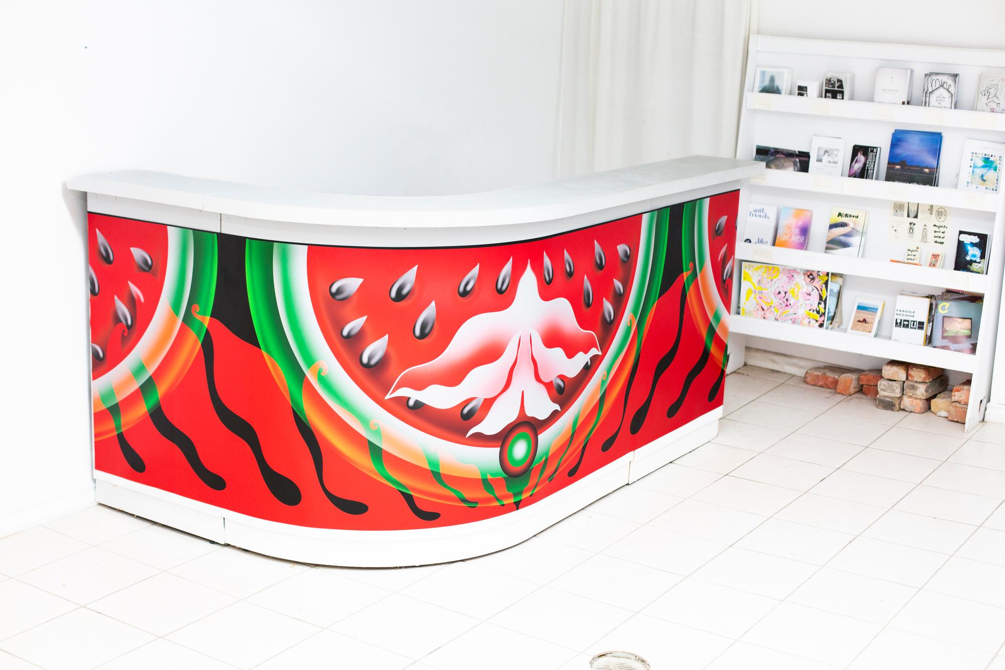 A gallery bar redecorated to look like an artwork in its own right through abstract compositions in green, red, and black, located next to a bookcase stakced with volumes.