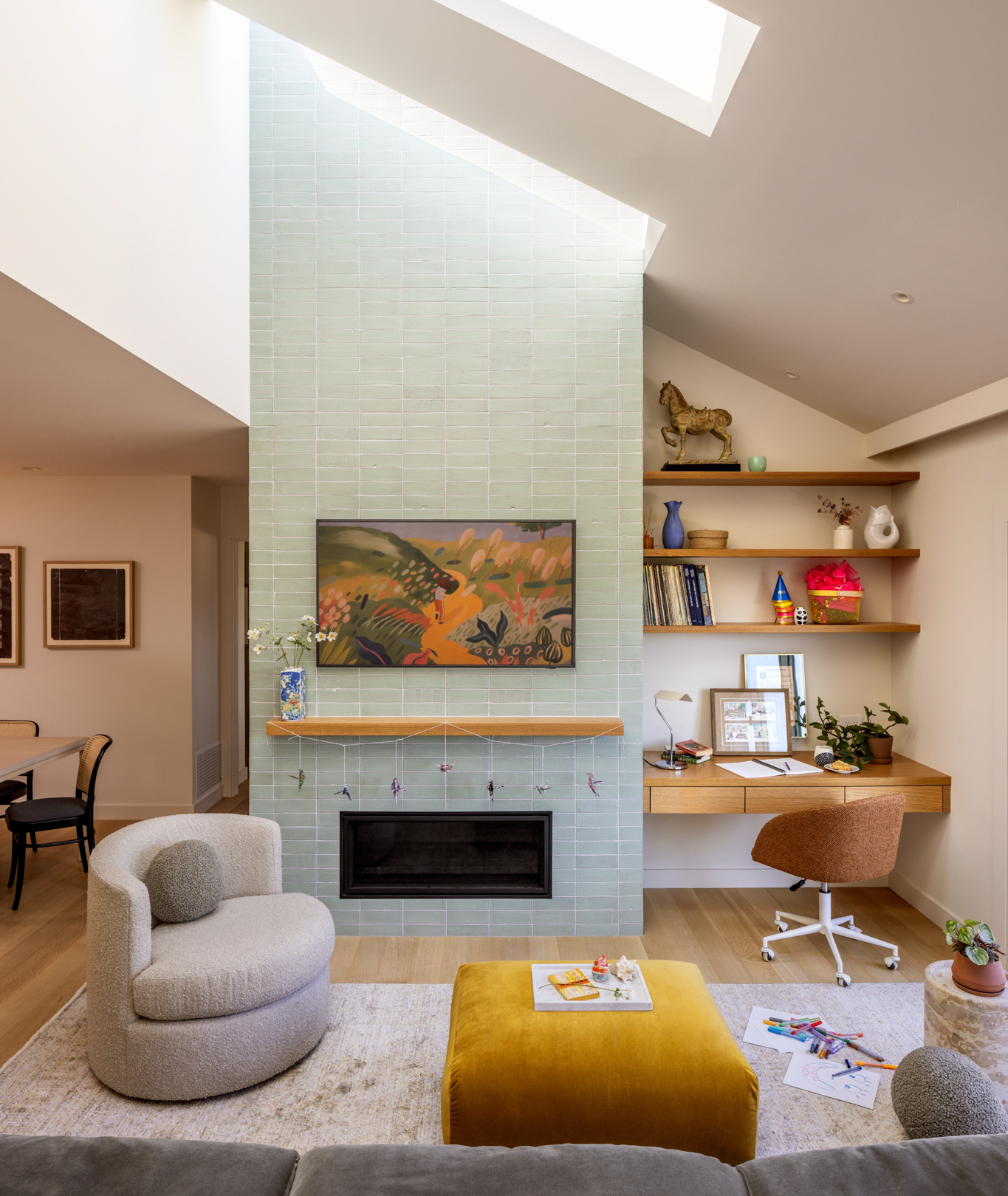 A modern, bright living space with a large skylight above, the chimney breast is clad in a mint green tile with a wooden beam mantle piece. There is a circular grey armchair and a mustard yellow ottoman.