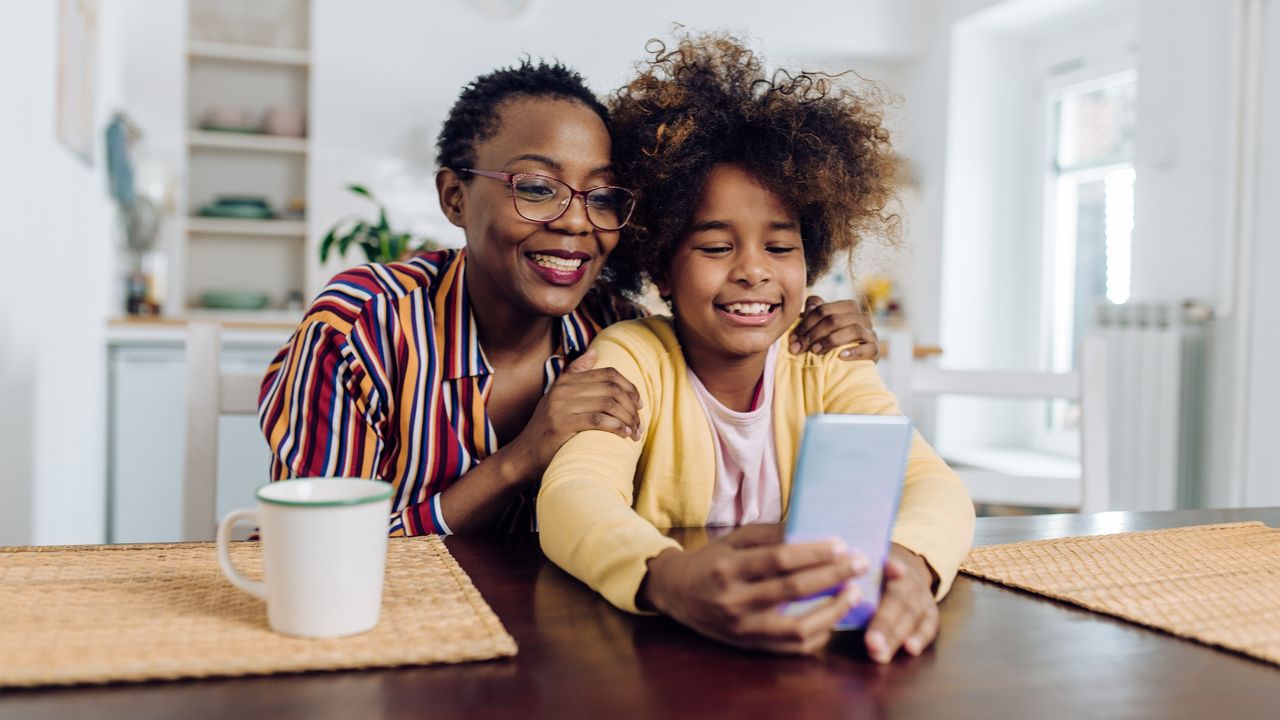 A mom and young daughter smile as they look at the daughter&#039;s phone.