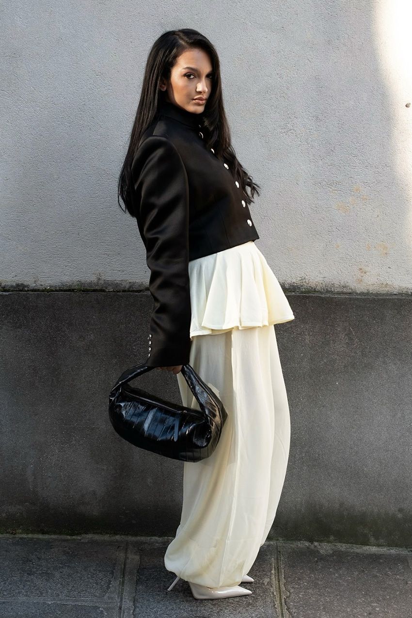 A street style photo of a woman standing outside in front of a gray wall wearing a black military jacket with white ballon pants, a black patent leather crescent bag, and pointed white pumps.