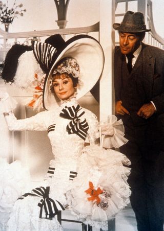 a promo shot of audrey hepburn wearing the white victorian gown and bit hat in my fair lady