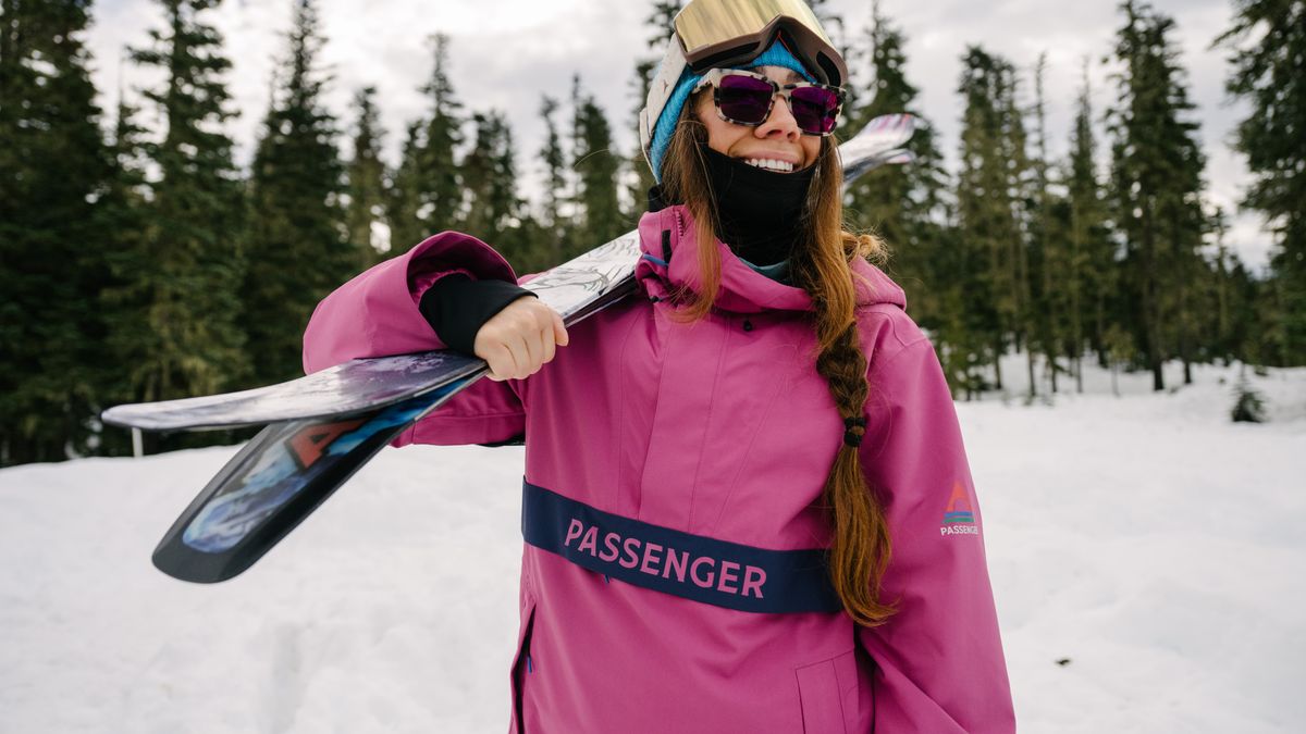 Woman skier in pink Passenger jacket holds skis over her shoulder