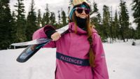 Woman skier in pink Passenger jacket holds skis over her shoulder