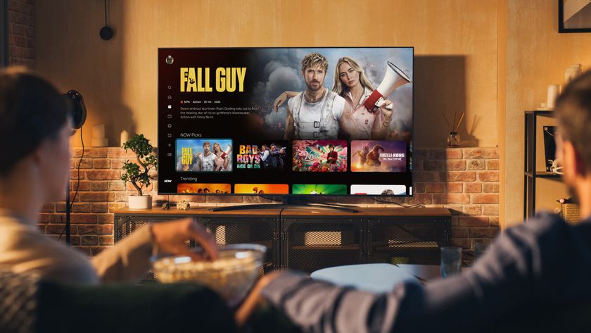 A TV showing the new Now home page with Free Guy as the featured selection. Two people sitting in the foreground watching the TV in a modern living room environment.