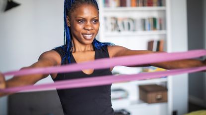 woman facing the camera holding a pink resistance band at both ends and pulling it apart. 