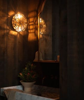 A dark bathroom with dark brown stained wood paneling, a circular wall sconce, a gold mirror on the wall and a marble sink with a plant on it