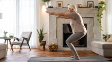 A woman performs shallow squats on an exercise mat in a living room.