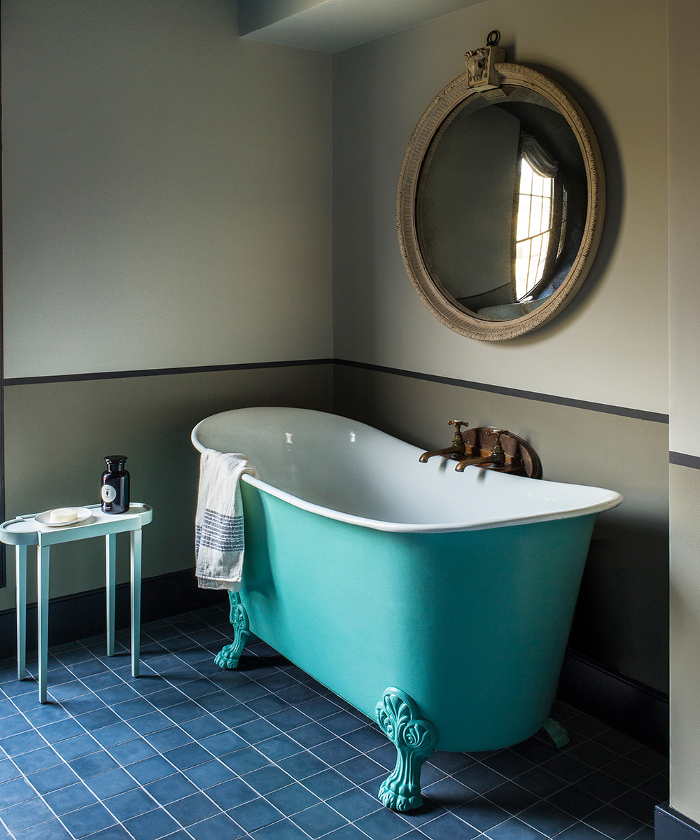 A turquoise free-standing bath in a bathroom with blue floor tiles and a round mirror on the wall