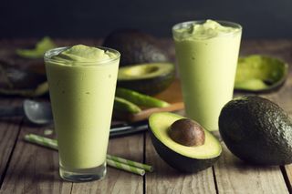 two avocado smoothies in glasses on a table surrounded by halved avocados for decoration