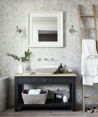 Intricate, gray tiled wall, square white mirror above a black and light wood sink unit, dark wood flooring, light wood towel ladder with white towels, two wall lamps either side of the mirror