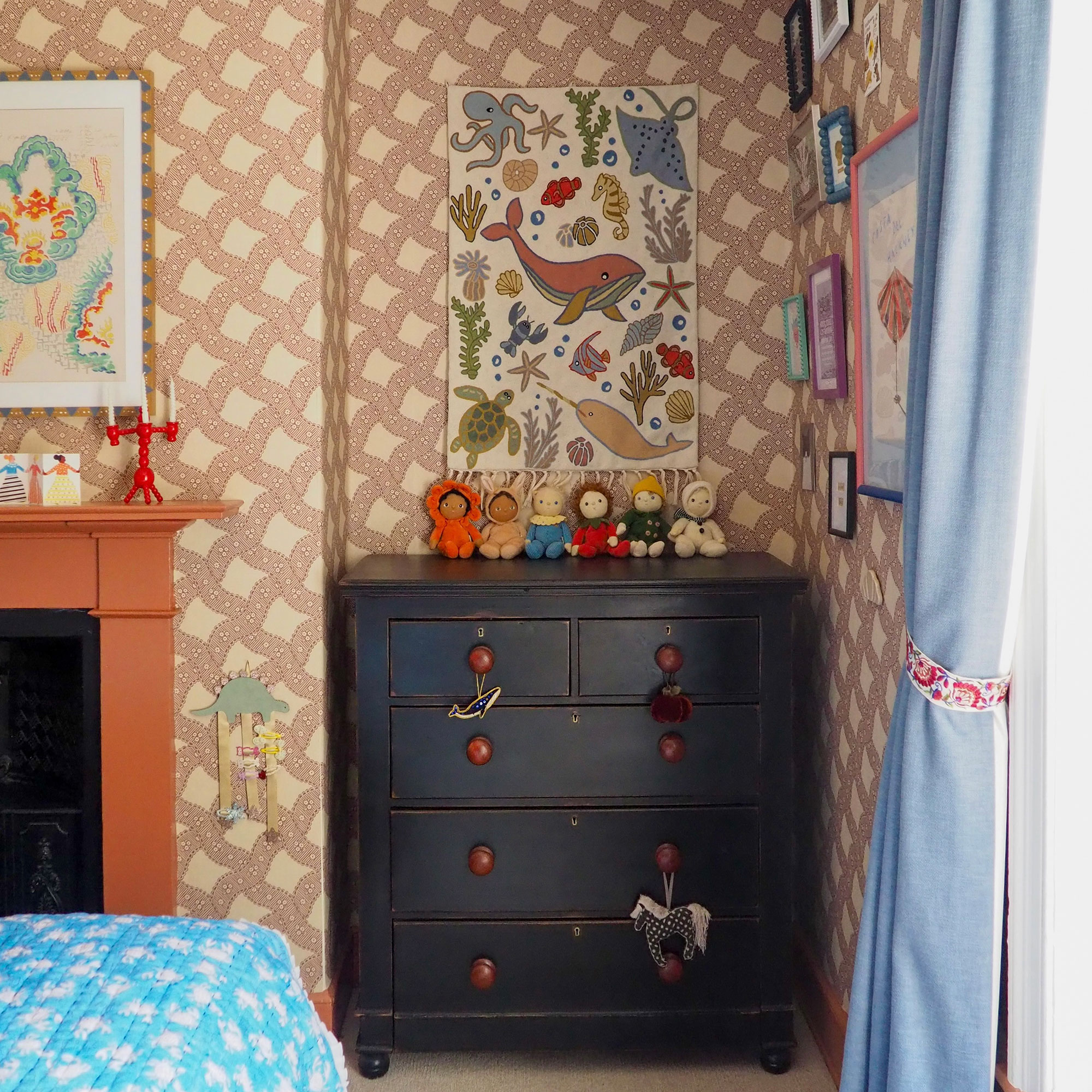 Child's bedroom with patterned wallpaper, dark chest of drawers, blue curtains and colourful wall hangings and prints