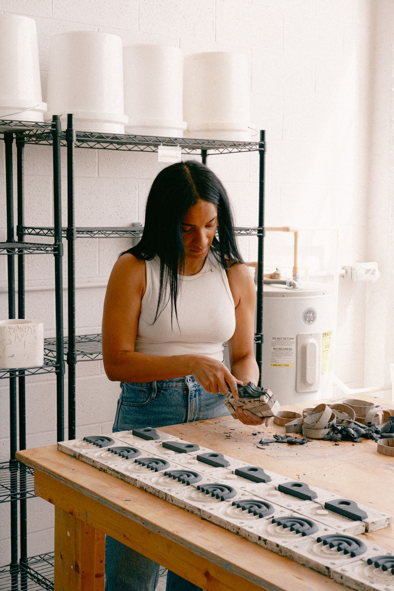 Images of a woman in jeans and a white top standing in a ceramic studio. There is a wooden table in front of her with several molds of mug handles. She is shaping one in the image.