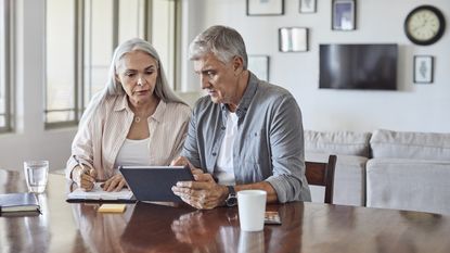 An older couple look serious as they look at their laptop on the kitchen table and go over bills.