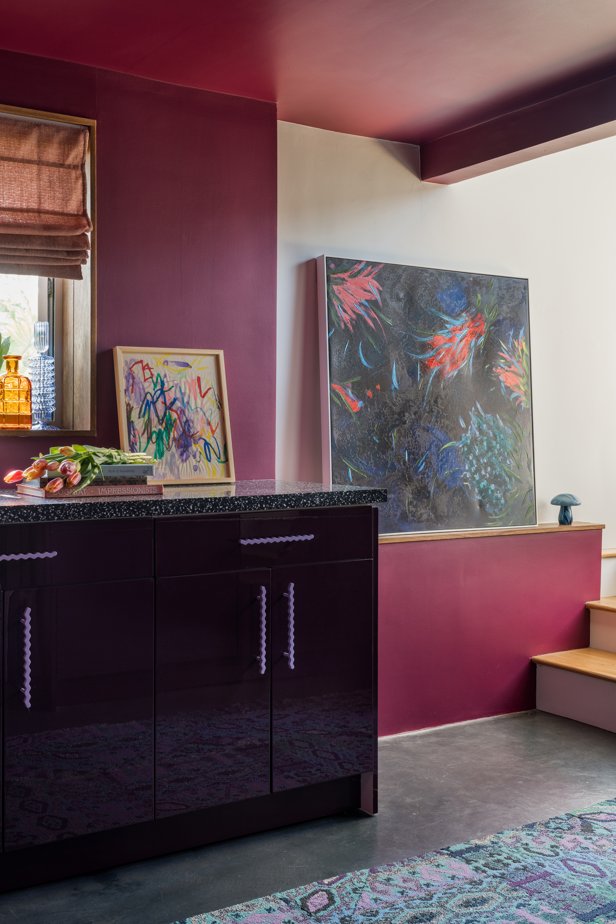 hallway with purple paint on ceiling and walls, large leaning artwork, black glossy console with purple handles styled with a bunch of flowers and small leaning artwork, polished concrete floors, and a blue, purple, green, and gray rug on floor