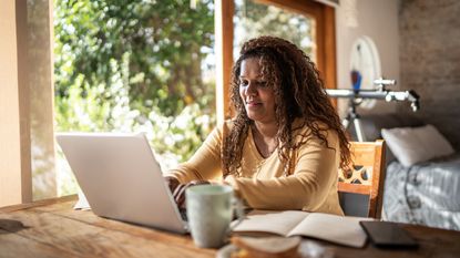 A woman smiles as she sits at her desk at home and looks at her laptop.