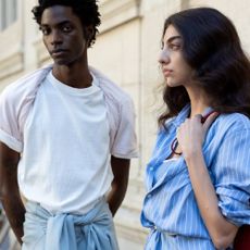 A male model wearing a white t-shirt from SSENCE, with a female model wearing a blue, pin-striped shirt from SSENSE. 