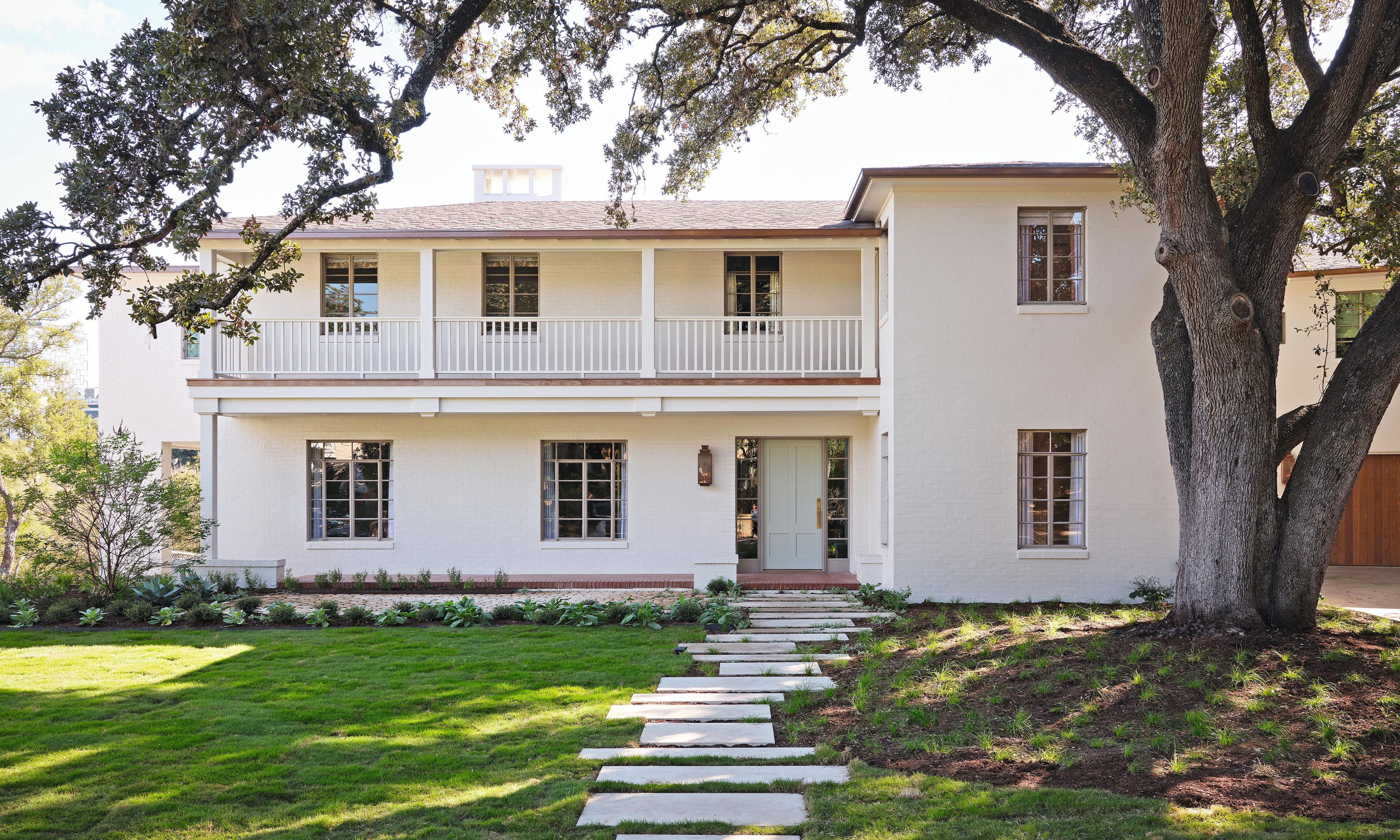 a white house in Austin with upper balcony, a large tree in the front yard with grass and stepping stones up to the front door